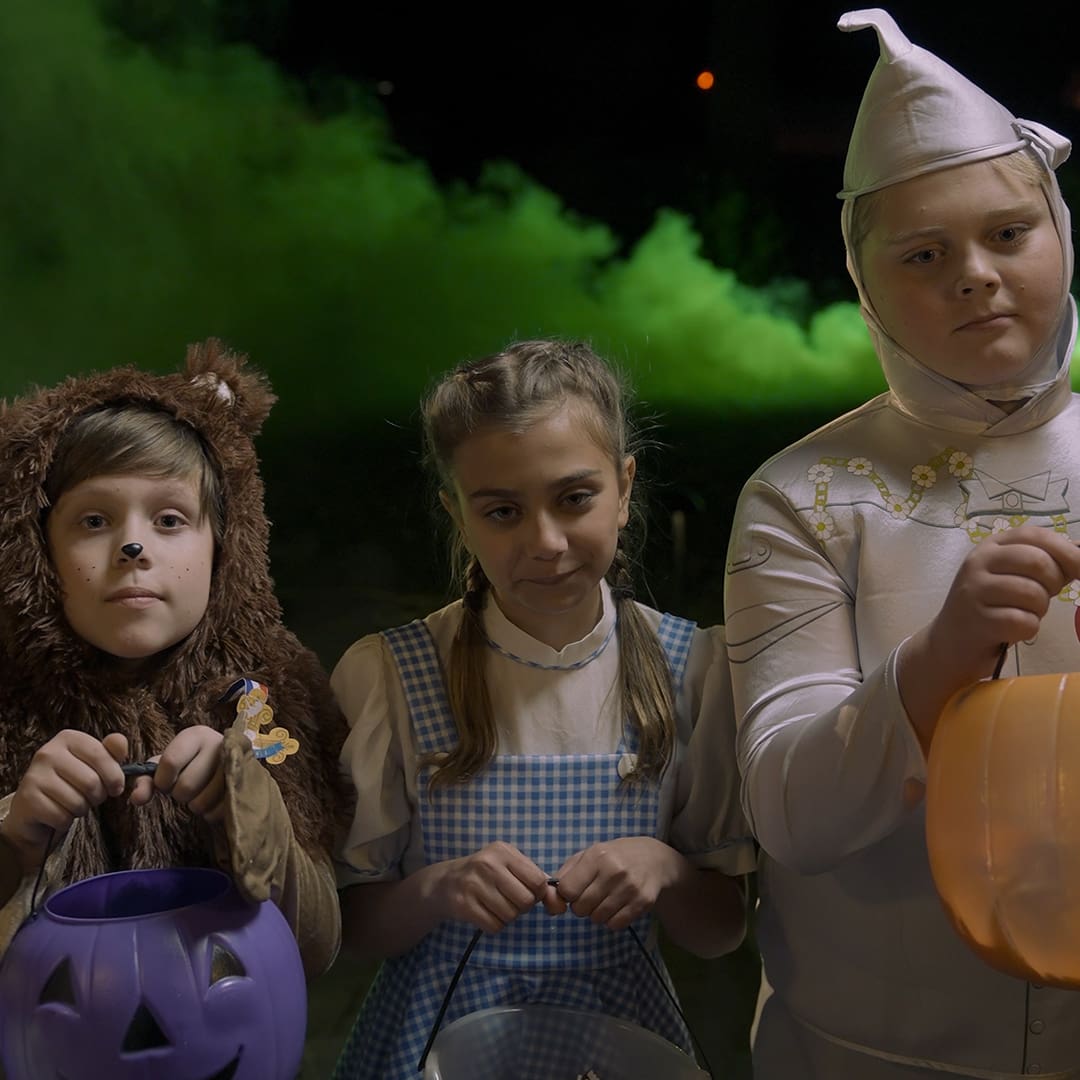 Three children in Halloween costumes stand outdoors at night. From left to right: a boy dressed as a brown bear, a girl dressed as Dorothy from The Wizard of Oz (in a blue and white gingham dress), and another child dressed as the Tin Man, holding an orange pumpkin bucket. A cloud of green smoke is visible in the background.