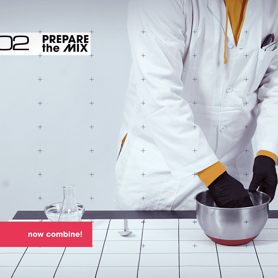 A person in a white lab coat and black gloves preparing a mix in a stainless steel bowl in a laboratory setting.