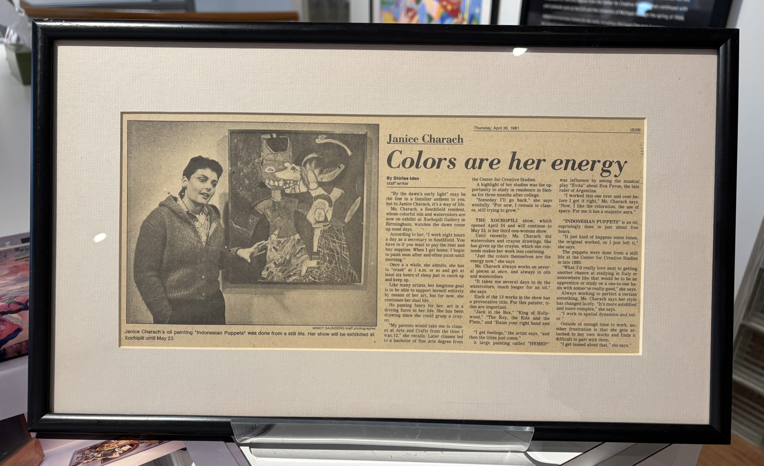 A framed, black-and-white newspaper article from 1981 titled "Colors are her energy." On the left, a photograph shows the artist Janice Charach standing next to her painting, "Indonesian Puppets," which is a vibrant, stylized depiction of three figures. The accompanying text details her art show and her painting style.