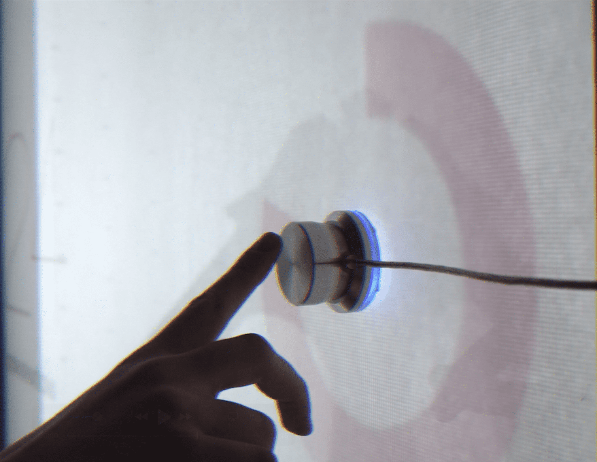 A close-up shot of a hand touching a small, cylindrical, clear physical knob or sensor mounted on a white, projected surface. The knob has a blue light emitting from its base, and a wire extends from its side. Faint red circular graphics are visible on the surface behind the device.