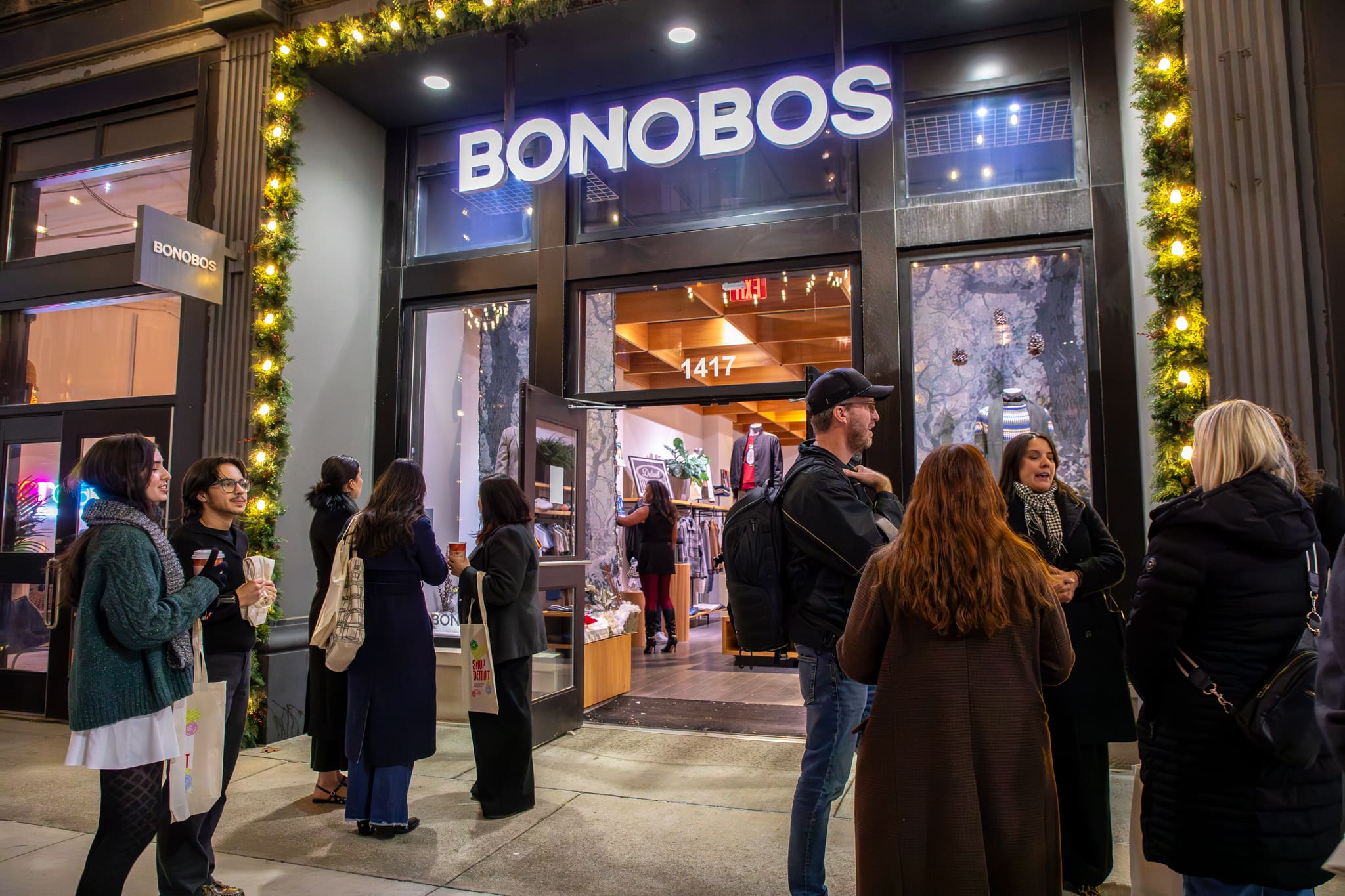 A group of people are gathered on a Detroit sidewalk at night in front of the Bonobos storefront.