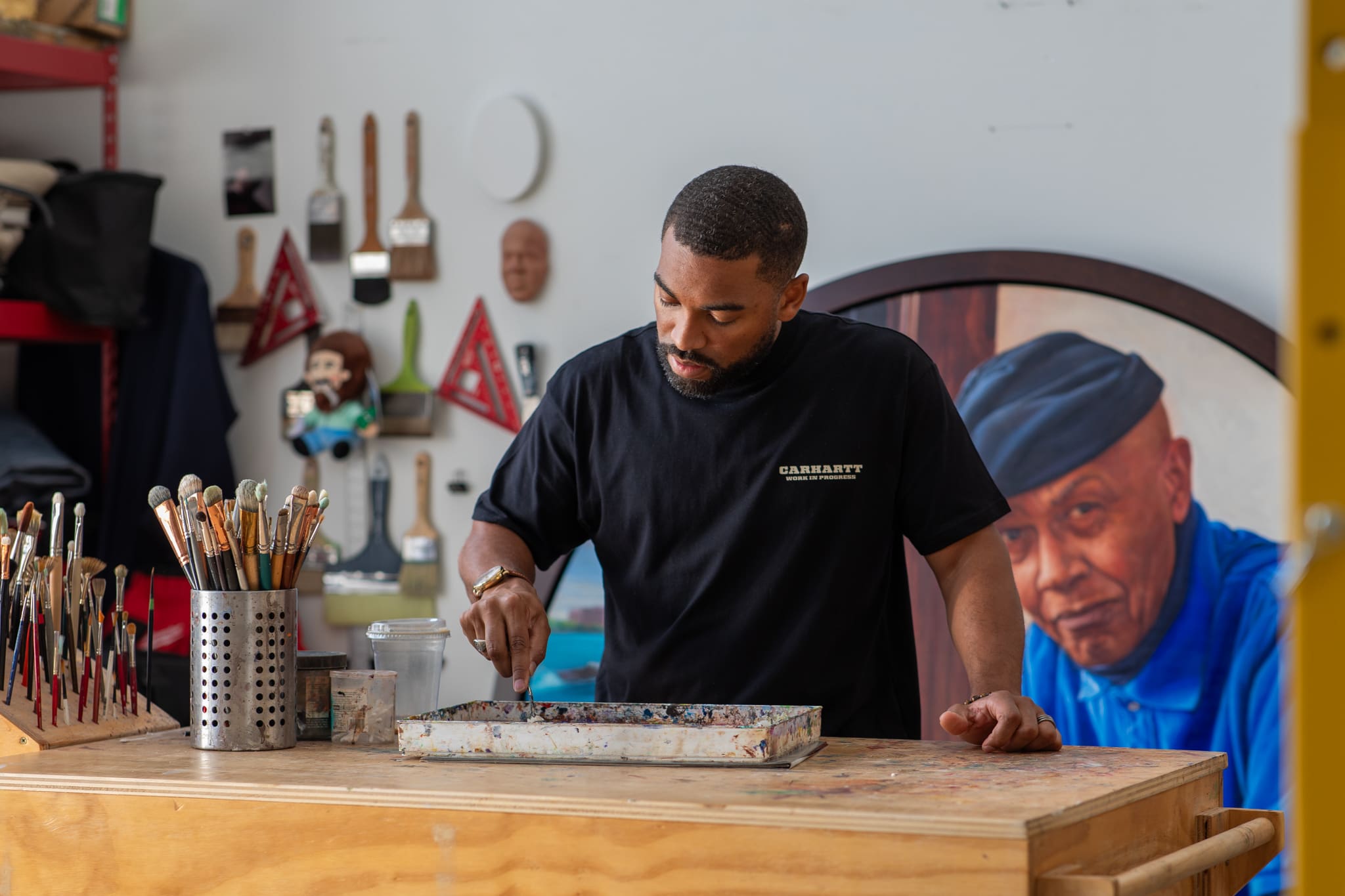 Artist Mario Moore stands at a wooden workbench in his Detroit studio, mixing paint on a palette. He is wearing a black Carhartt t-shirt and looking down intently at his work. The studio walls are lined with brushes, tools, and a large circular portrait of a man in a blue shirt.