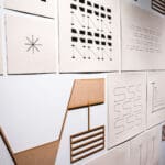 Close-up angled view of a wall installation featuring square paper prints with geometric typography and abstract laser-cut wooden shapes.