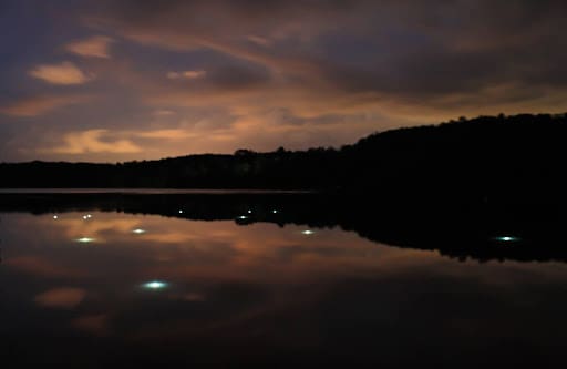 A dark, secluded lake at night reflects a cloudy, softly illuminated sky. Small points of white light are submerged beneath the water’s surface, scattered across the lake to mirror a constellation, creating a calm, immersive scene where the glowing lights appear both above in the sky and below in the water.