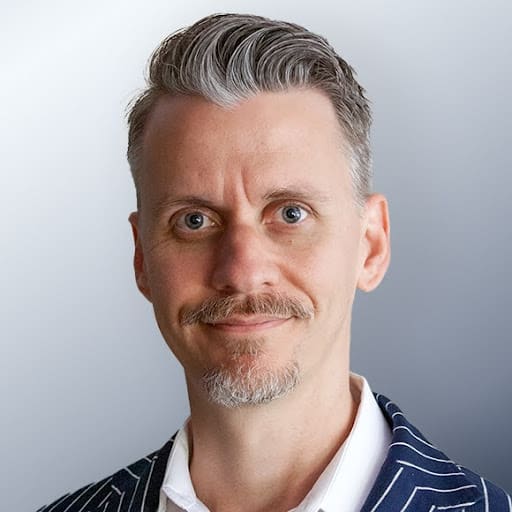 Headshot of a man with short gray hair and a trimmed goatee, wearing a white shirt and a dark pinstriped jacket, against a light gray background