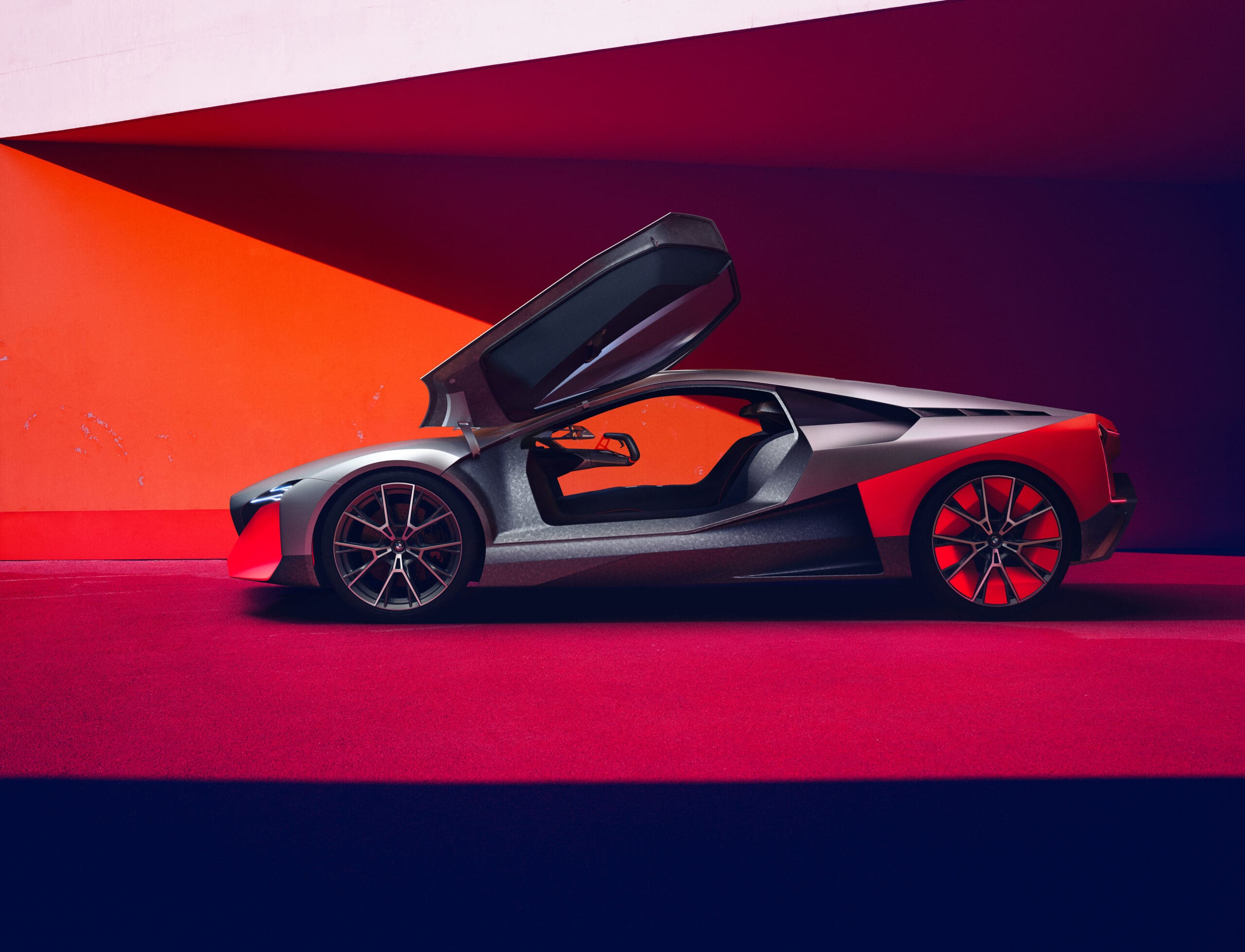 A profile view of the BMW Vision M NEXT concept car with its butterfly door open, featuring a silver and neon orange color scheme against a matching geometric background.