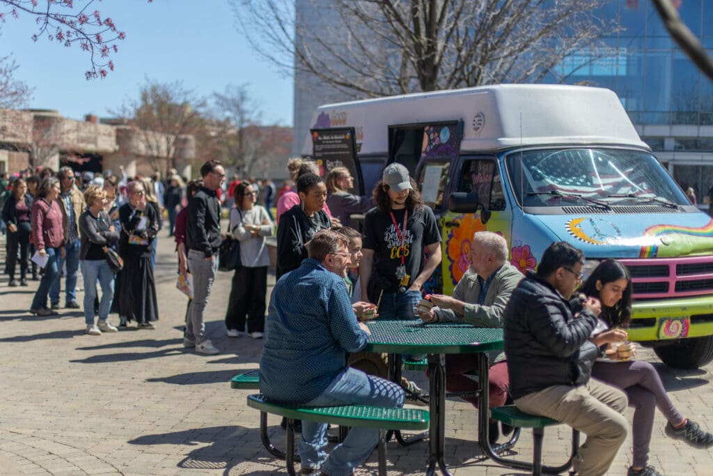 A bright, sunny outdoor scene at the College for Creative Studies. A long line of students stands on a brick plaza waiting at a colorful, tie-dye decorated food van featuring a rainbow and peace sign. In the foreground, a diverse group of students and faculty sit at a green circular picnic table, eating and talking.