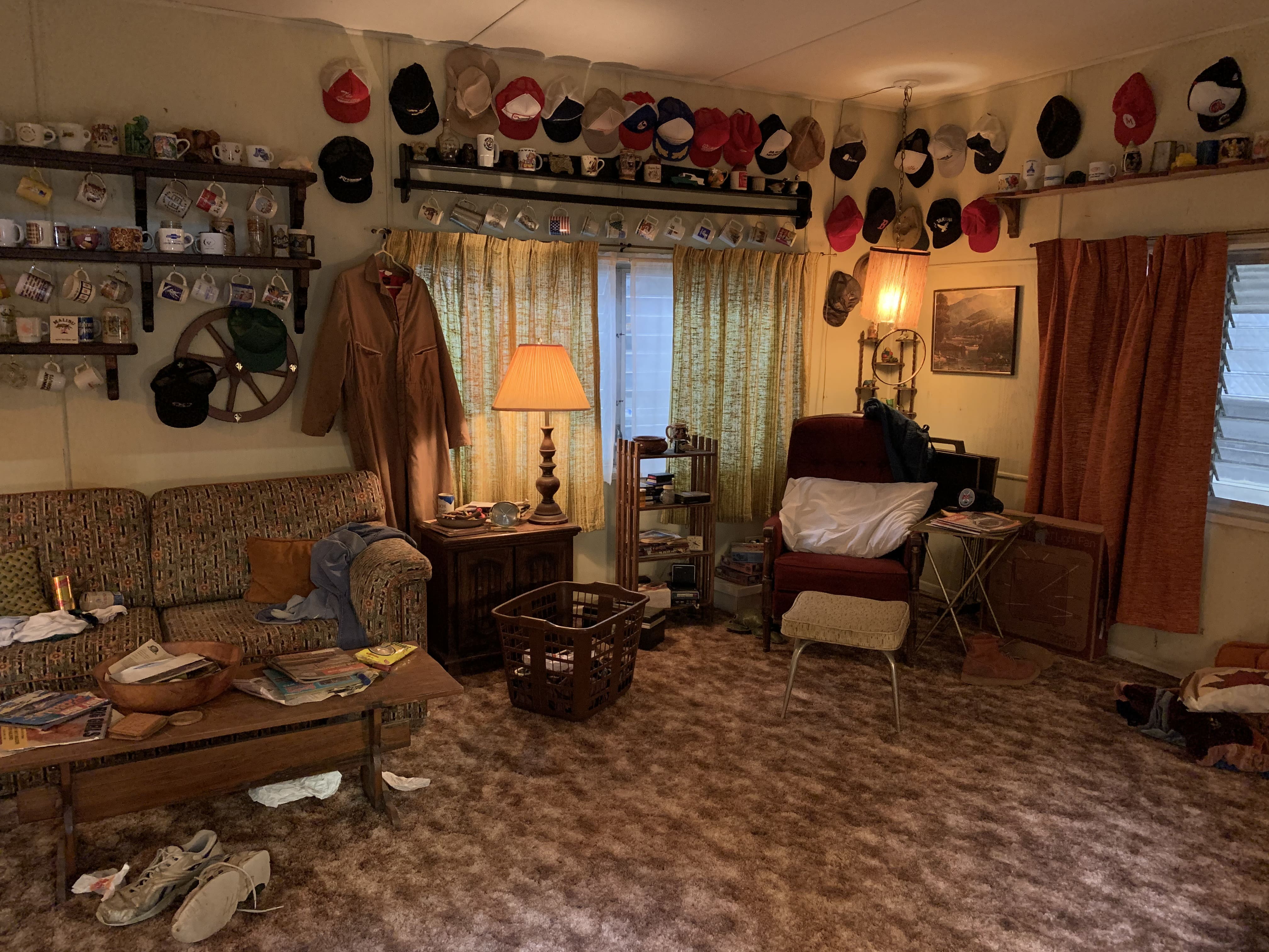 A highly detailed 1980s trailer living room set from Stranger Things. The room features wood-paneled walls decorated with dozens of trucker hats and coffee mugs. A messy, lived-in space with brown shag carpet, a vintage floral couch, and 80s-era decor, lit by warm indoor lamps.