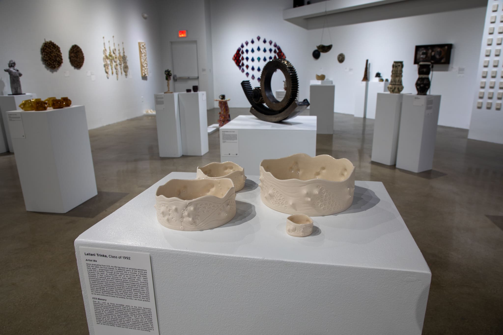 Four off-white, textured ceramic bowls of varying sizes sit on a white pedestal in the foreground of a brightly lit art gallery. In the background, other sculptural ceramic works are displayed on pedestals and walls, including a large dark circular gear-like piece. An artist bio for Leilani Trinka, Class of 1992, is attached to the side of the pedestal.