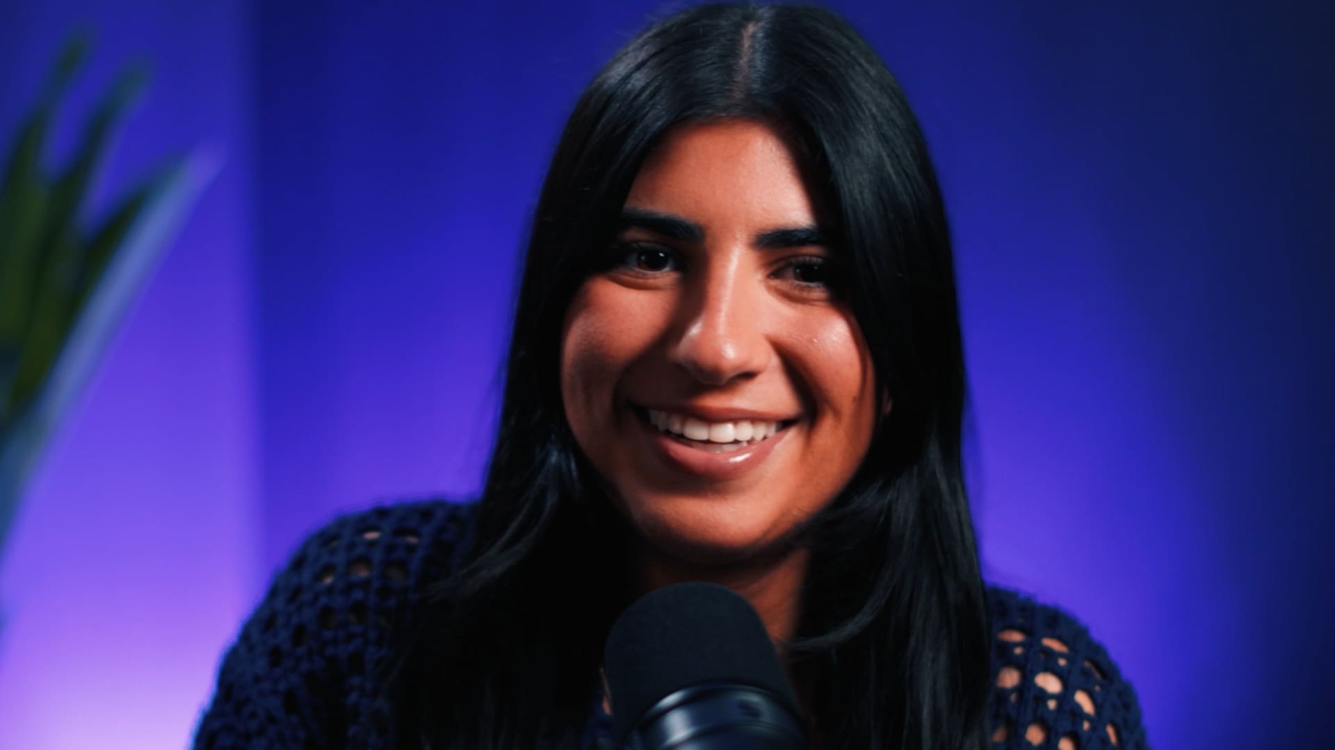 A vibrant, close-up shot of Annabelle Saco smiling during a podcast or interview recording. She is positioned behind a professional black studio microphone. The background is a striking, out-of-focus deep blue and purple gradient, with the blurred leaves of a plant visible on the left side. She is wearing a dark, textured knit sweater.