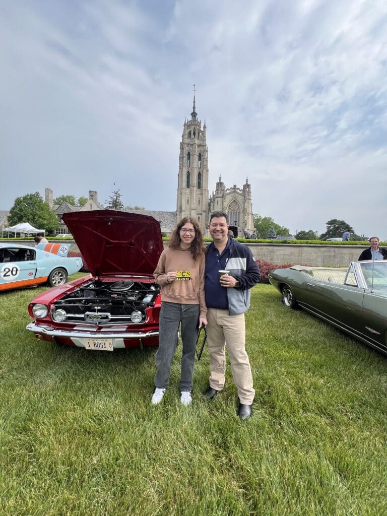 A young woman wearing a brown "CCS Alumni" sweatshirt and a man smiling together on a grassy field at an outdoor car show. Behind them is a classic red Ford Mustang with its hood open, revealing the engine. To the left, a light blue race car with orange stripes is partially visible, and to the right is a vintage olive green convertible. In the background, a large, ornate Gothic-style stone cathedral with a tall spire rises against a cloudy sky.