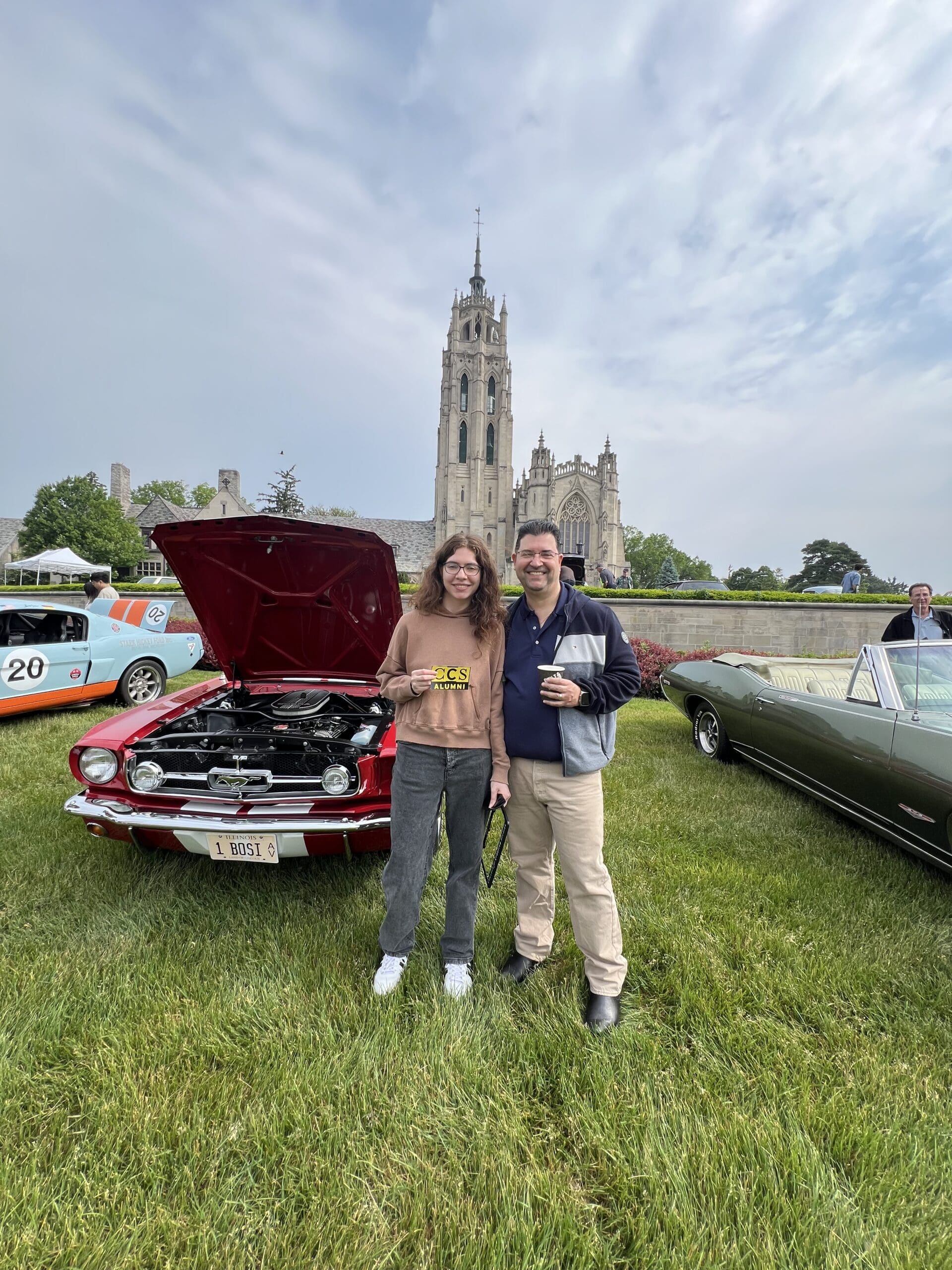 A young woman wearing a brown "CCS Alumni" sweatshirt and a man smiling together on a grassy field at an outdoor car show. Behind them is a classic red Ford Mustang with its hood open, revealing the engine. To the left, a light blue race car with orange stripes is partially visible, and to the right is a vintage olive green convertible. In the background, a large, ornate Gothic-style stone cathedral with a tall spire rises against a cloudy sky.
