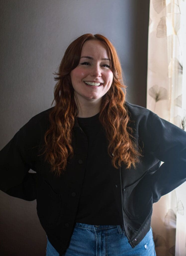 A smiling woman with wavy red hair and a black jacket posing confidently against a dark wall in soft, natural window light.