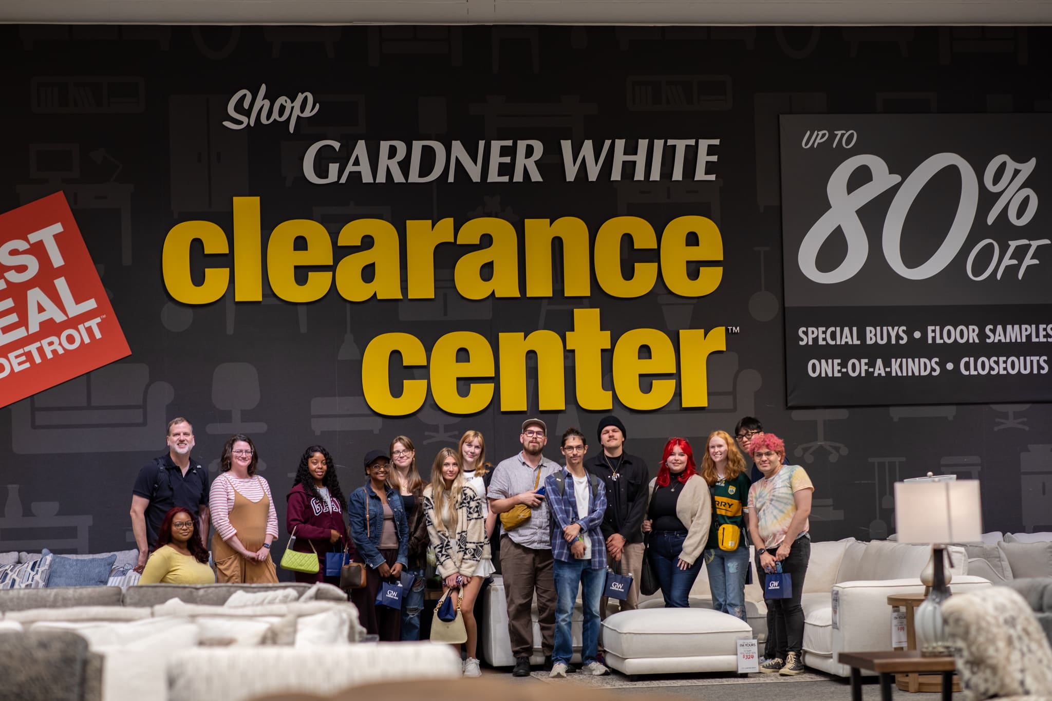 A group of 14 students and faculty pose in front of a massive black and yellow 