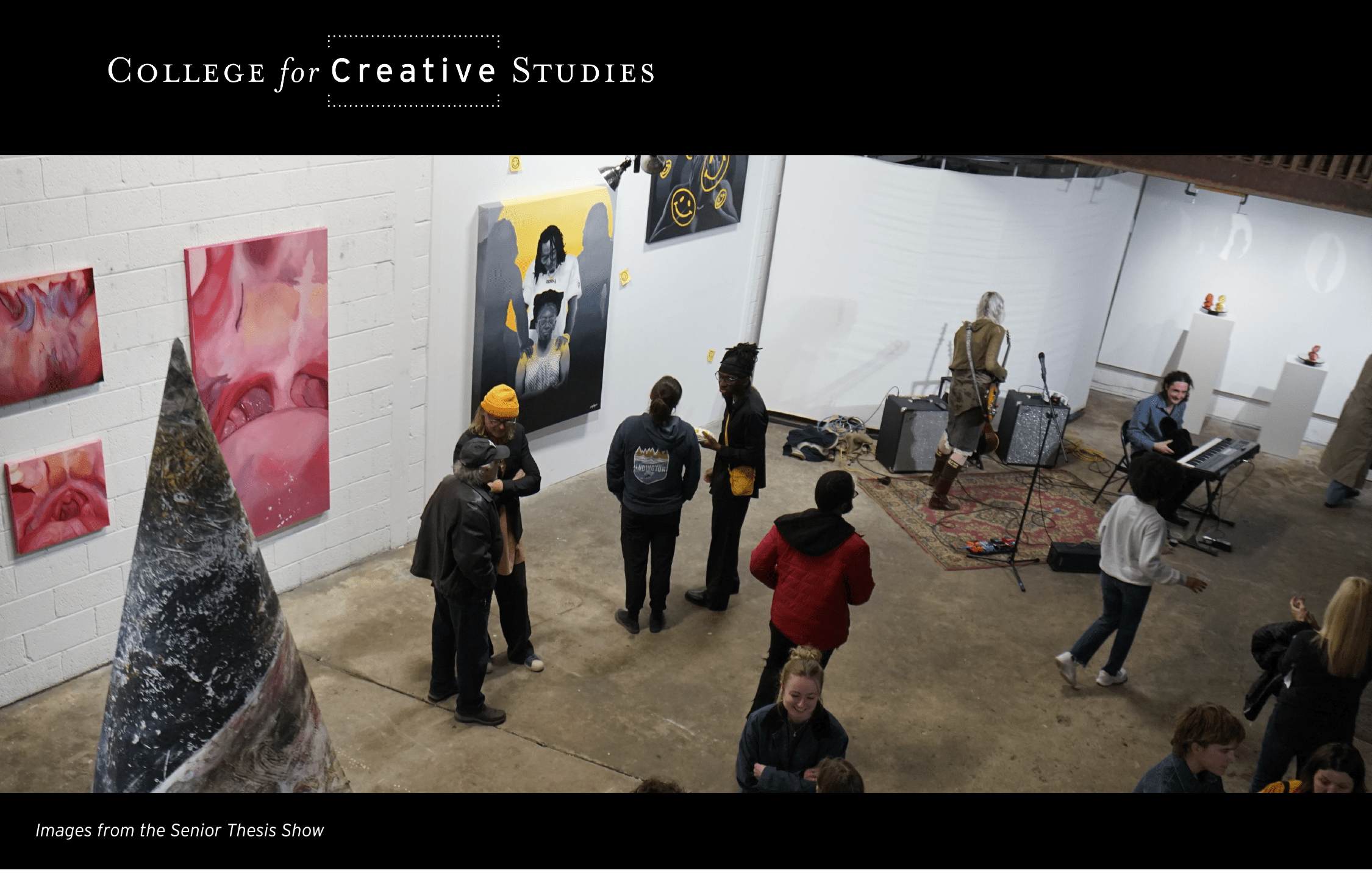 High-angle view of the College for Creative Studies Senior Thesis Show. Students and visitors gather in an industrial gallery space featuring large-scale paintings, a tall cone-shaped sculpture, and a live band performing on a rug.
