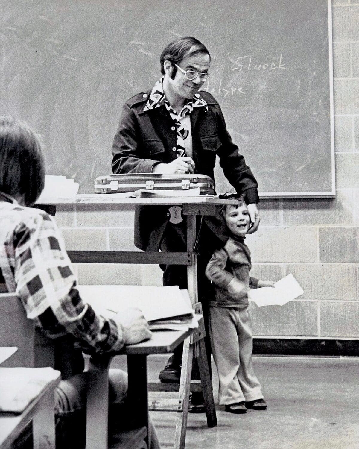 Professor James S. Goldwasser with his young son at the College for Creative Studies (CCS) in the 1970s. This candid black-and-white photography student's snapshot captures a heartwarming moment during a design lecture, featuring the professor at a drafting table and his laughing child.
