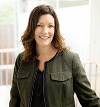 A professional headshot of Shannon McPartlon, a woman with shoulder-length wavy brown hair, smiling and wearing an olive green wool jacket with black leather trim.