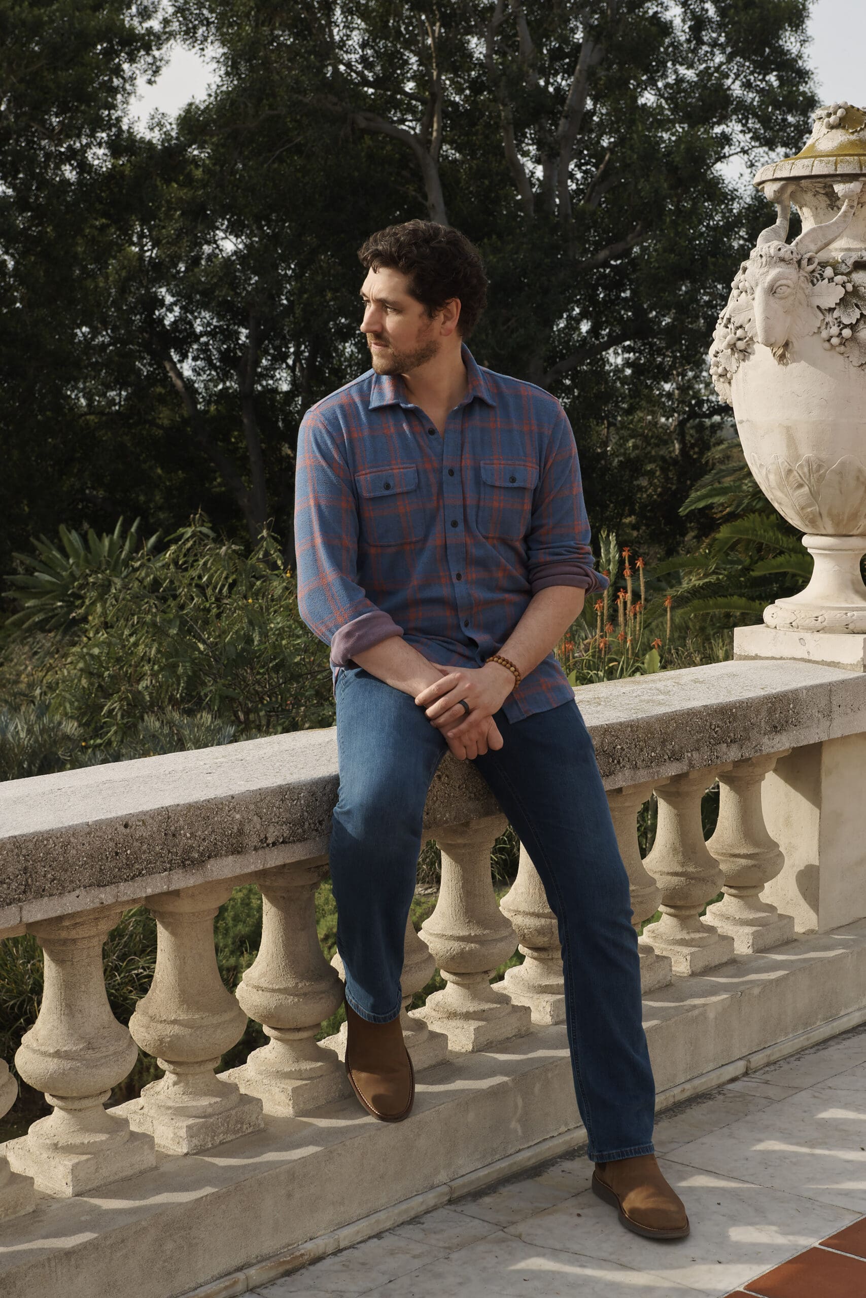 260129_943 A full-length portrait of Michael Maher Jr. smiling with his arms crossed on a path at the Huntington Gardens. He is wearing a blue and orange plaid shirt and jeans. He is surrounded by a variety of desert plants, including large succulents and cacti, with tall trees and autumn leaves in the background.