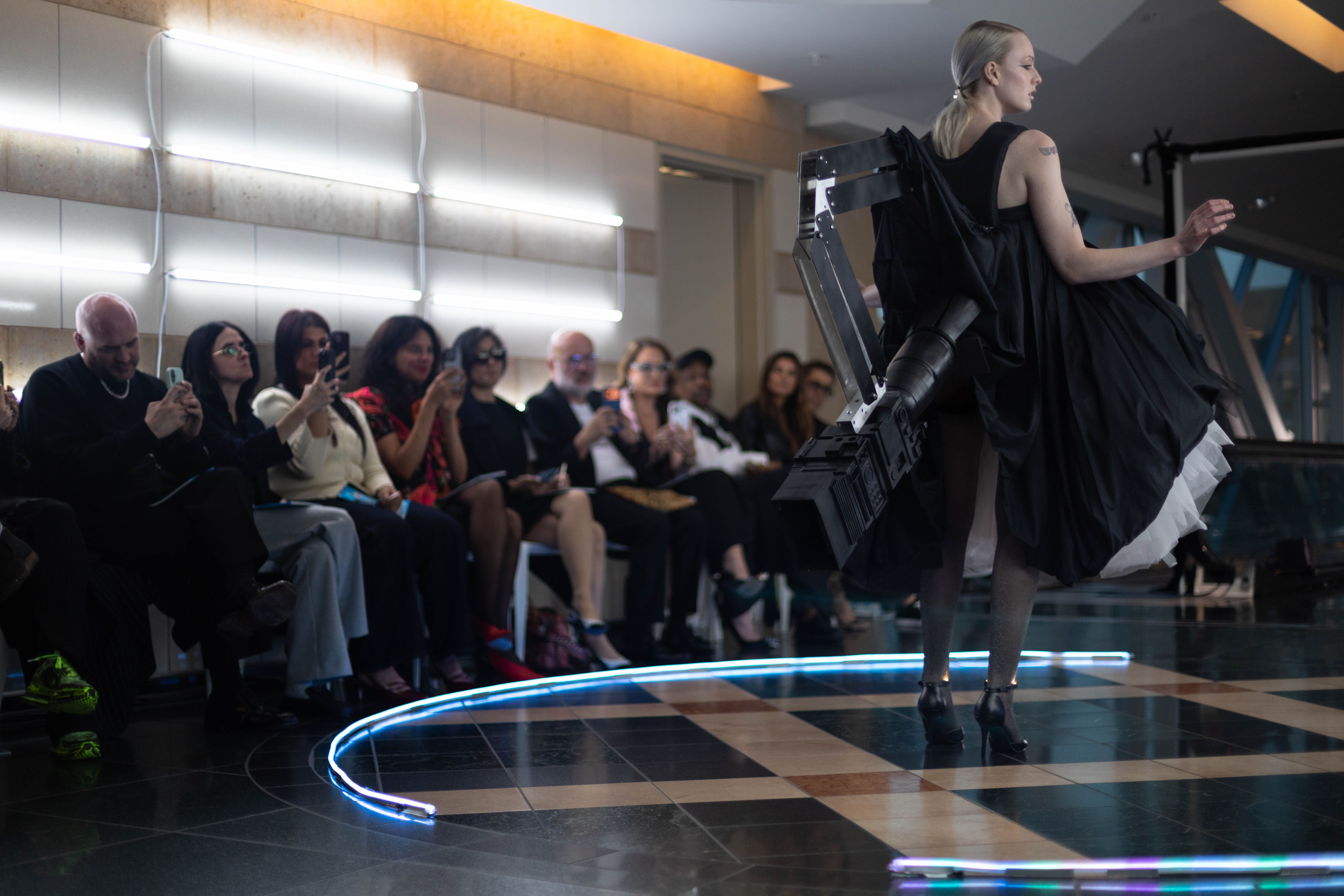 A model showcases a student design by Quade Feller on a circular runway during a Fashion Design Jury presentation. The avant-garde black dress features a large, mechanical industrial backpack. Jury members Danialle Karmanos, Millie de la Valette, Diana Costescu, Silvia Prada, and Stuart McMillan sit in the background taking photos under modern neon lighting.