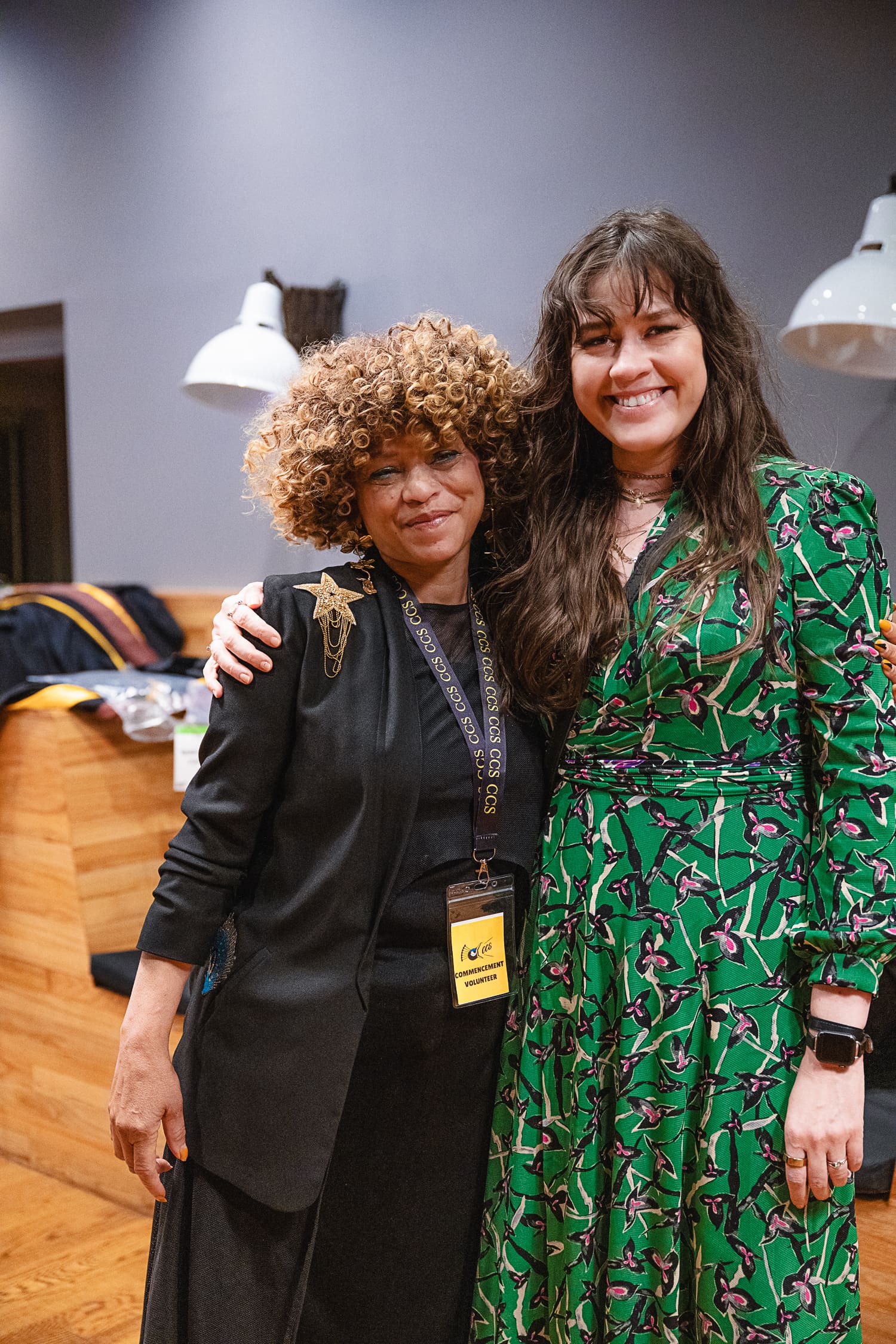 A medium shot of Sabrina Nelson and Veronika Scott posing closely together at the CCS Commencement ceremony. Sabrina, on the left, has a voluminous blonde afro and wears a black blazer with a gold star pin and a volunteer lanyard. Veronika, on the right, has long brown hair with bangs and wears a bright green floral wrap dress. Both are smiling warmly at the camera in a brightly lit indoor space.