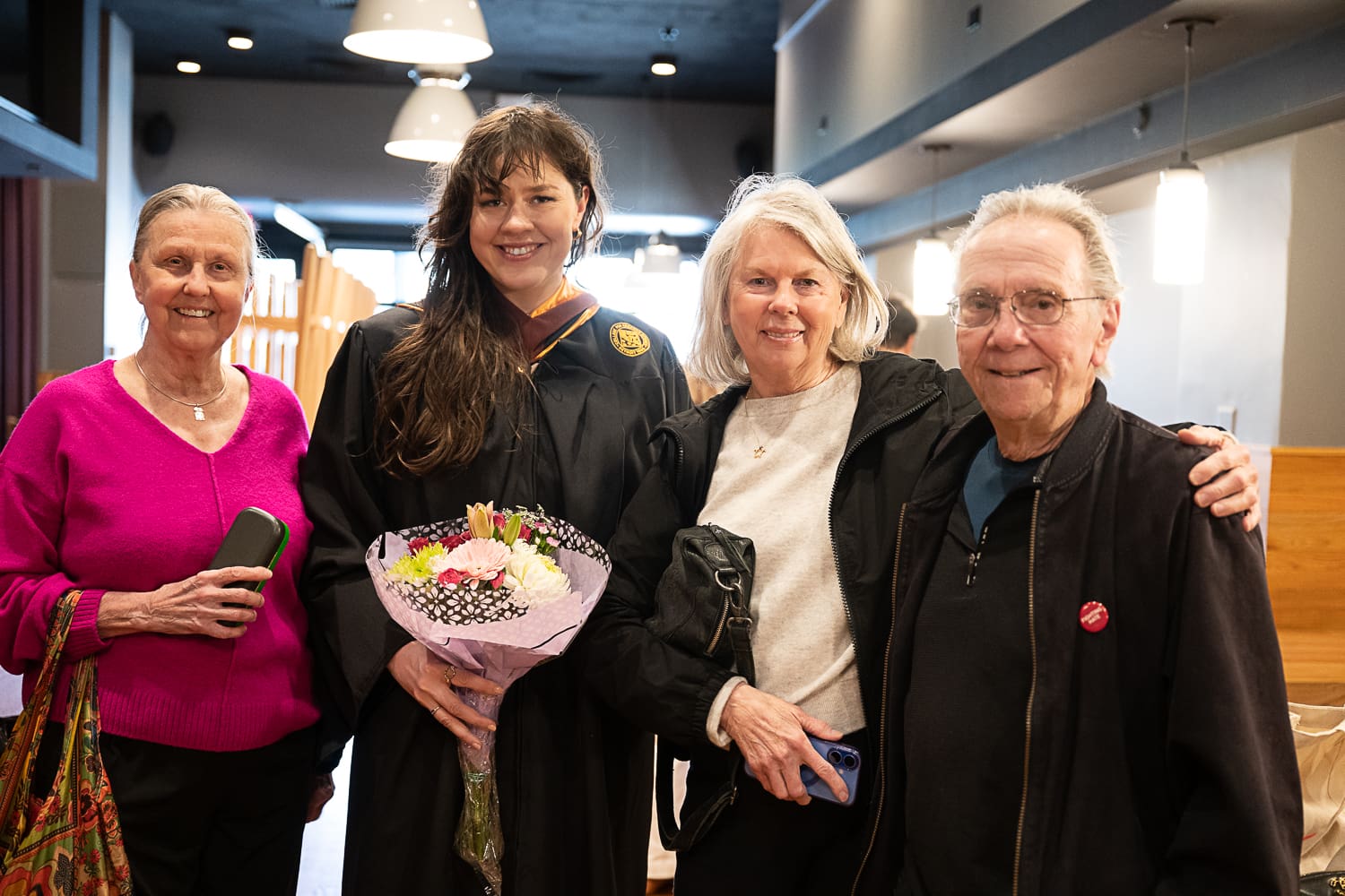 Veronika Scott, wearing a black graduation gown and gold stole, stands at the center of a group of three family members at the CCS Commencement ceremony. Veronika holds a bouquet of pink, white, and yellow flowers. To her left, a woman wears a bright magenta sweater; to her right, a woman in a black jacket and a man in a black zip-up sweater and glasses smile for the camera. They are gathered in a modern indoor venue with warm lighting.