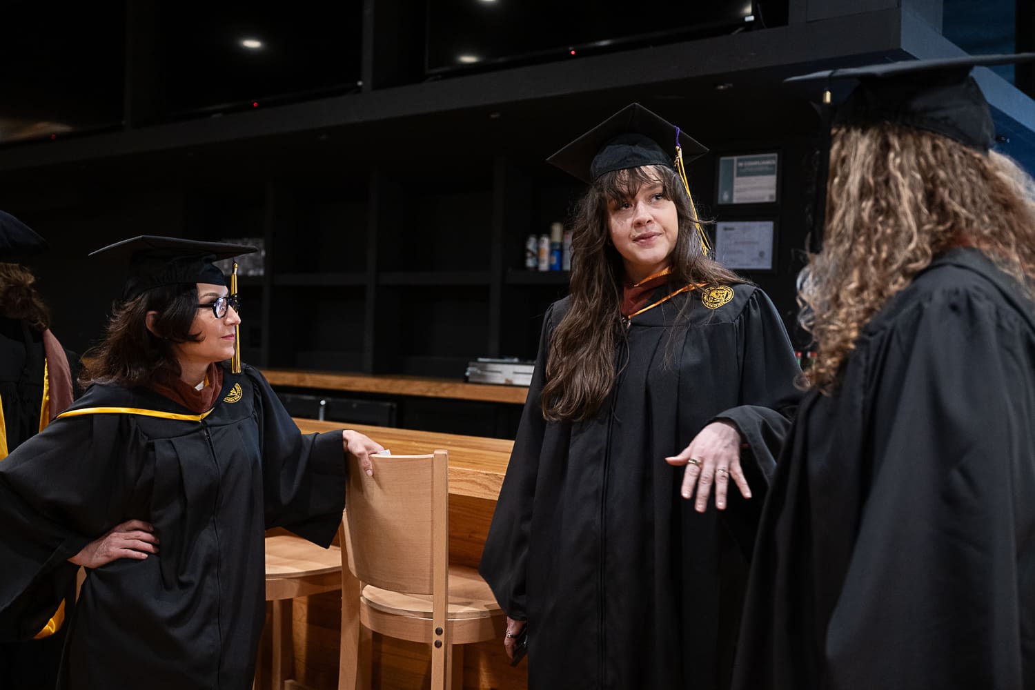Three women in black graduation gowns and caps converse indoors. At the center, Veronika Scott looks toward Denice Thomas (right), who has long curly hair. To the left, Darcel Deneau, wearing glasses, leans against a wooden counter while listening. The scene is candid, set against a dark background with a wooden bar-style table in a modern venue.