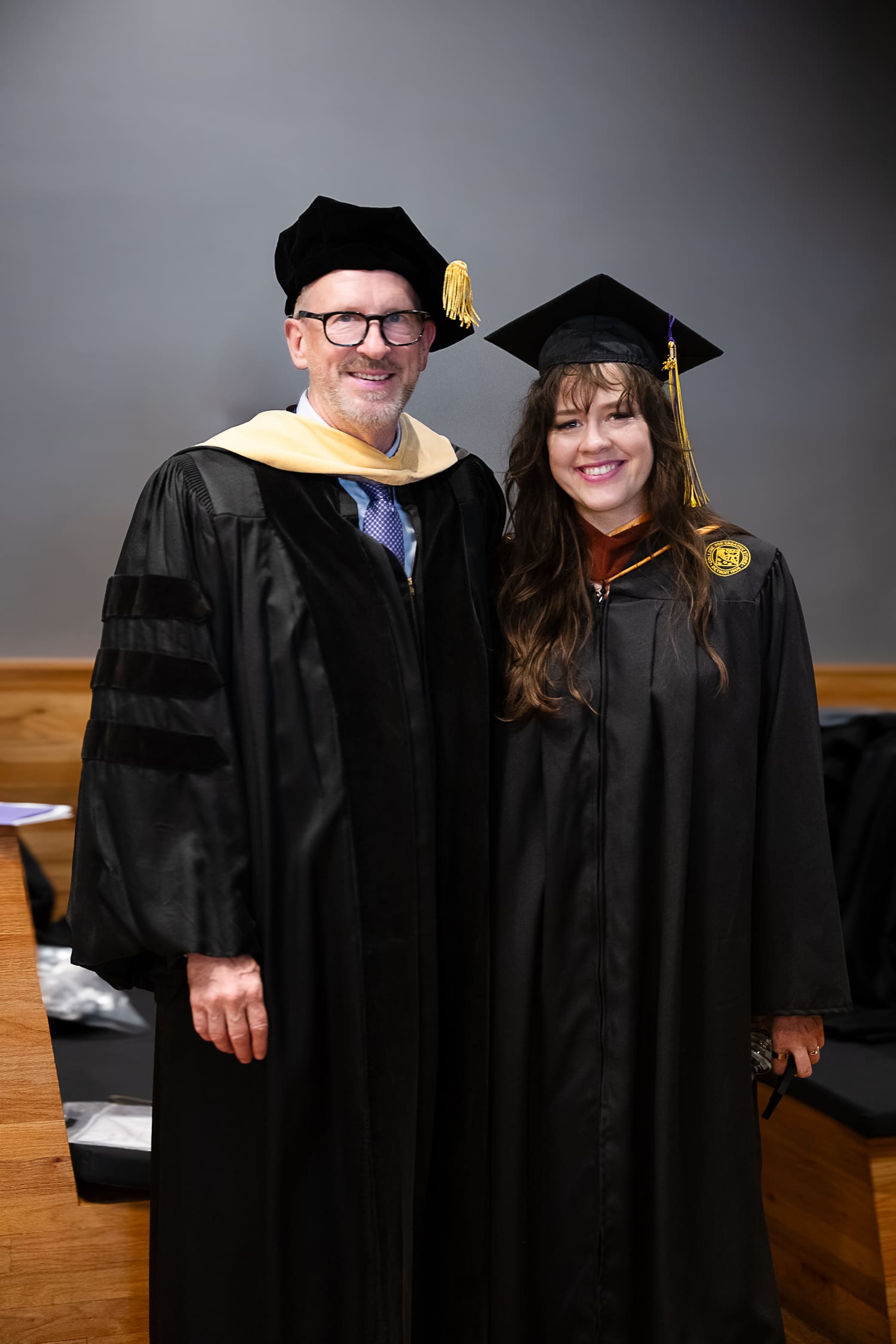 President Don Tuski and Veronika Scott stand side-by-side in their graduation regalia at the CCS Commencement ceremony. On the left, Don Tuski wears a doctoral gown with velvet bars on the sleeves, a yellow hood, and a black velvet tam. On the right, Veronika Scott wears a black master's gown with a gold stole and a traditional square mortarboard cap with a yellow tassel. Both are smiling at the camera against a neutral grey wall.