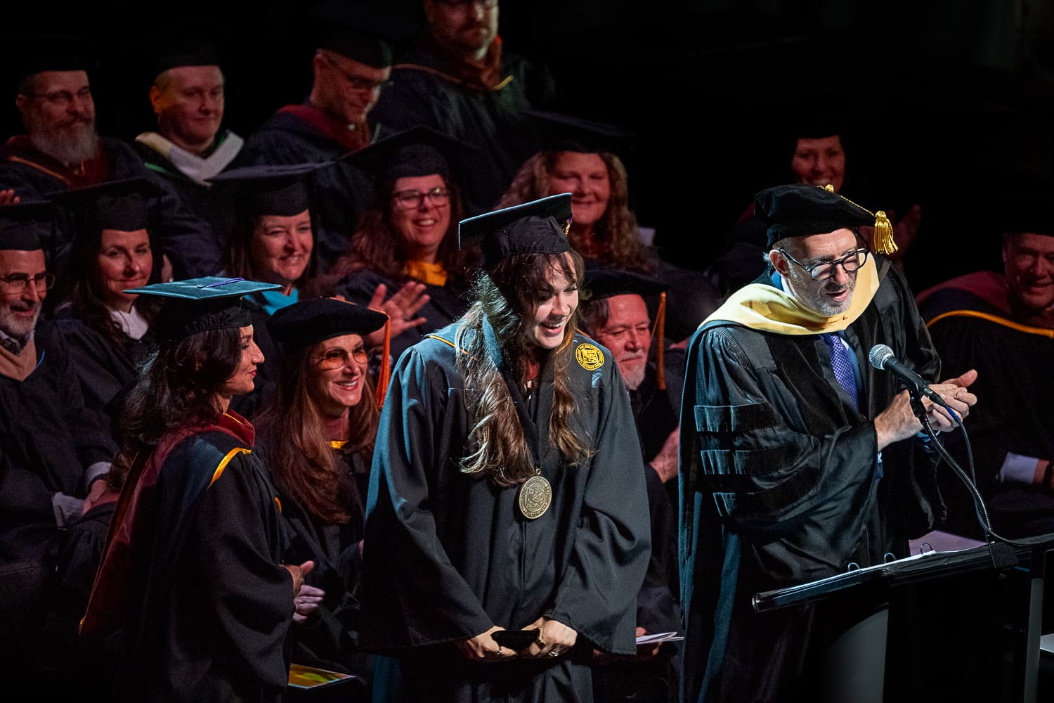 A medium shot on stage at the CCS Commencement ceremony. Veronika Scott stands at the center, smiling joyfully and wearing a large gold medallion over her graduation gown. To her right, President Don Tuski speaks into a microphone at the podium, gesturing toward her. In the background, faculty members and graduates in academic regalia are seated and clapping. The scene is warmly lit by spotlights against a dark theater background.