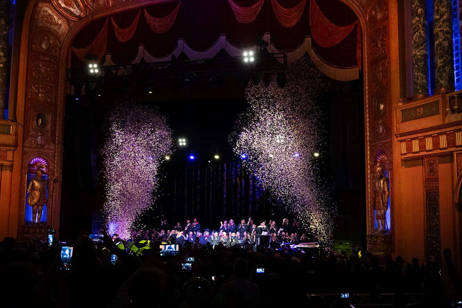 A wide, grand-scale shot of an ornate theater during the CCS Commencement finale. Two massive plumes of silver confetti erupt from the stage, soaring toward the high ceiling. On stage, a group of graduates and faculty are silhouetted against blue and white stage lights. The theater features elaborate gold-leaf arches, red velvet curtains, and large classical statues flanking the stage. In the darkened foreground, the silhouettes of audience members hold up glowing smartphones to capture the moment.