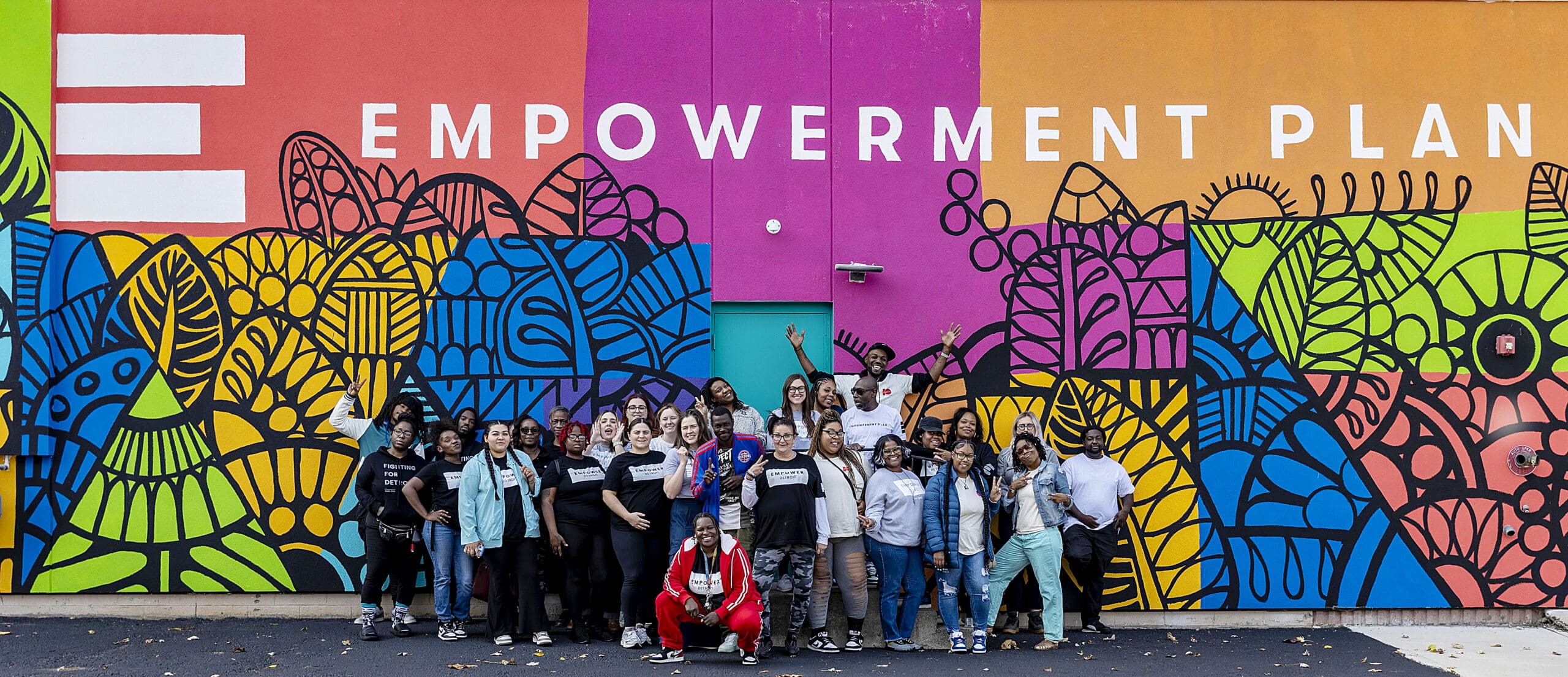 A wide panoramic shot of Veronika Scott and a large, diverse team of over twenty people posing in front of a massive, colorful mural at the Empowerment Plan headquarters. The mural features bold blocks of orange, pink, and yellow with intricate black line art of leaves and patterns, and the words 