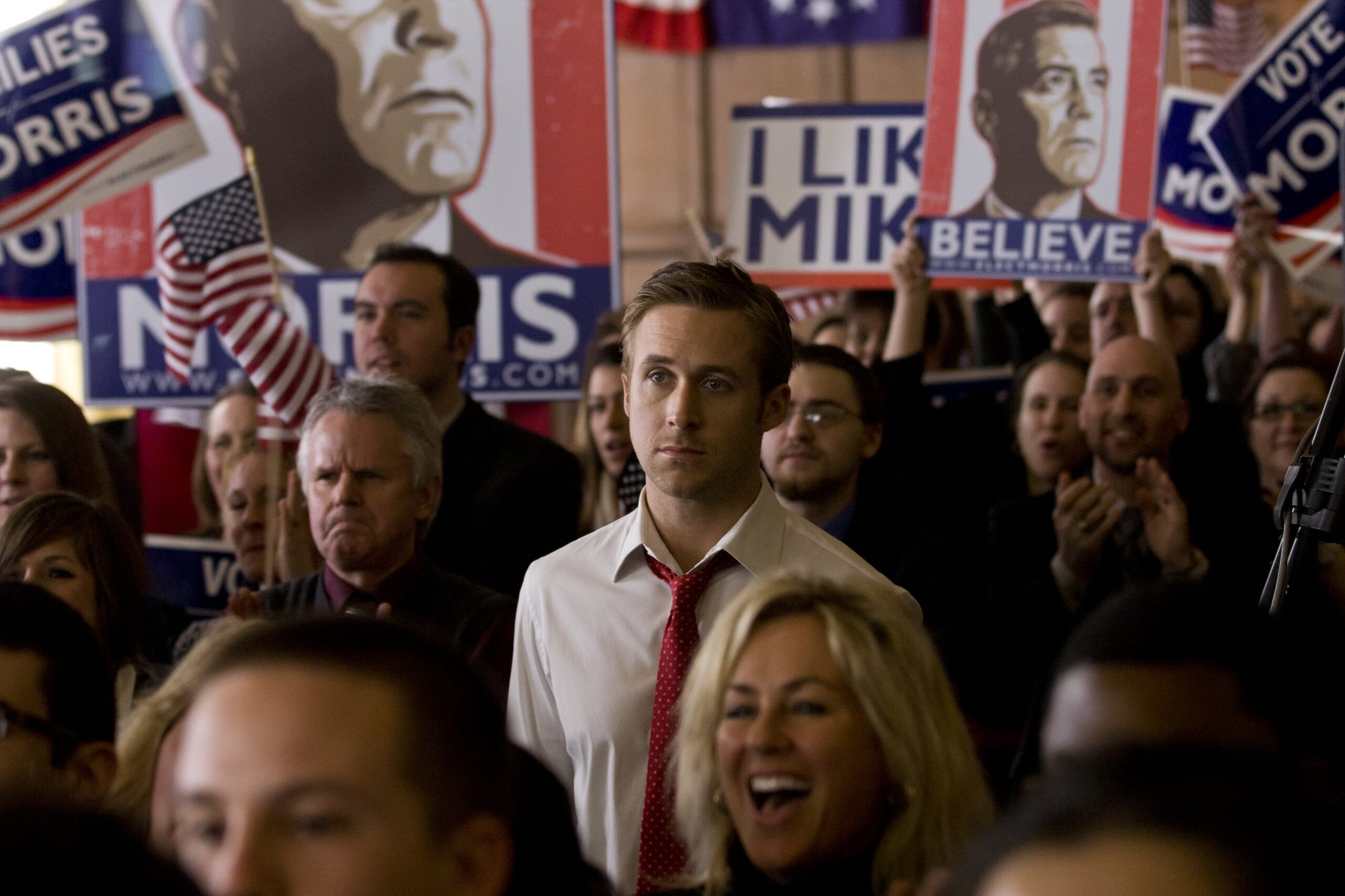 prop posters by Michael Maher Jr A medium shot of Ryan Gosling as Stephen Meyers in "The Ides of March," standing stoically in a crowded campaign rally. Behind him, supporters hold various political prop posters designed by Michael Maher Jr., featuring George Clooney's character, Mike Morris, with slogans like "Believe" and "I Like Mike."