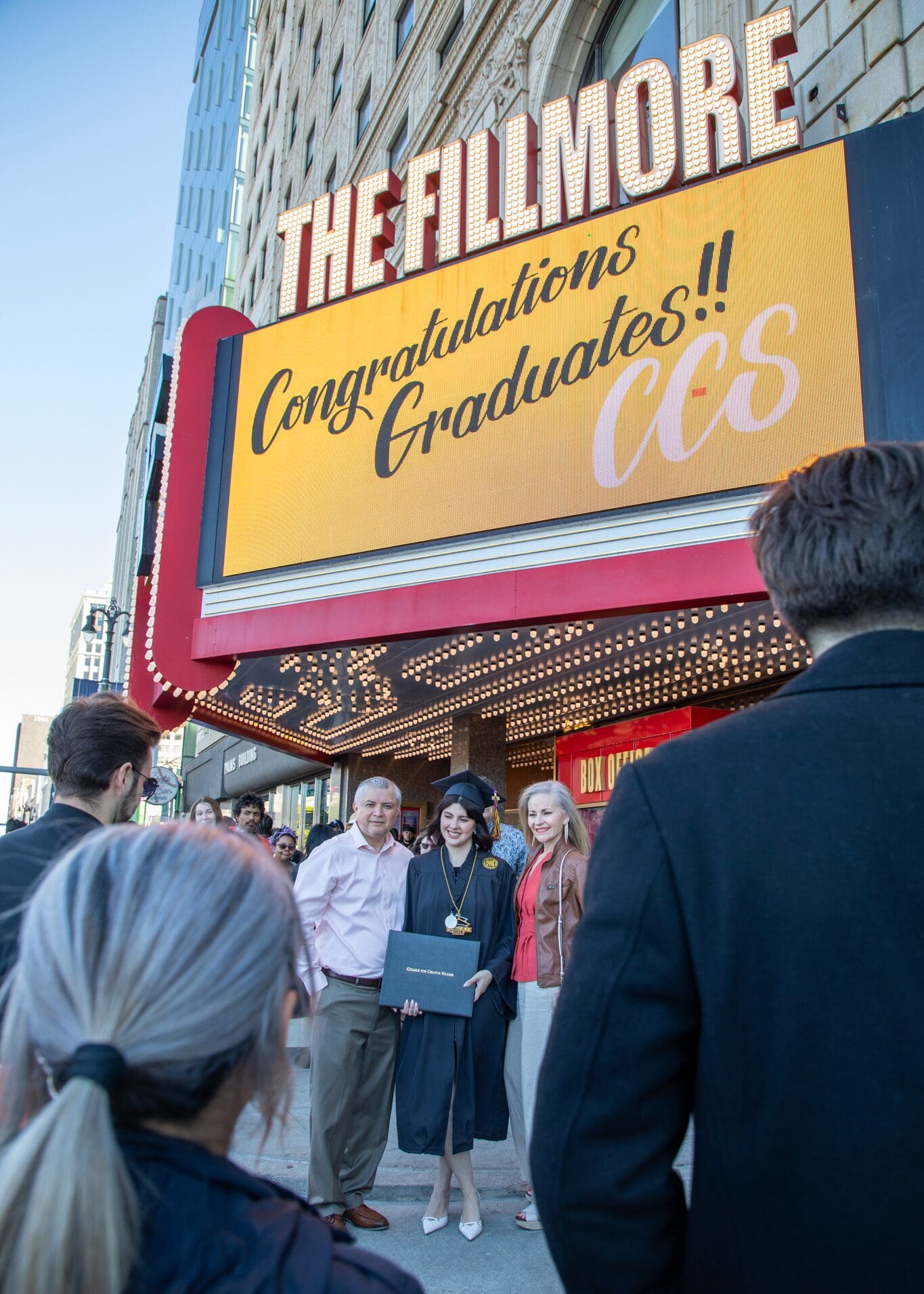 View from behind onlookers of a smiling graduate in a cap and gown posing with two family members on the sidewalk beneath a large, lit marquee sign that reads 'The Fillmore: Congratulations Graduates!! CCS'