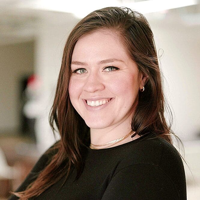 Veronika Scott is pictured with long dark hair in a black shirt, posing with arms crossed in a bright office setting.
