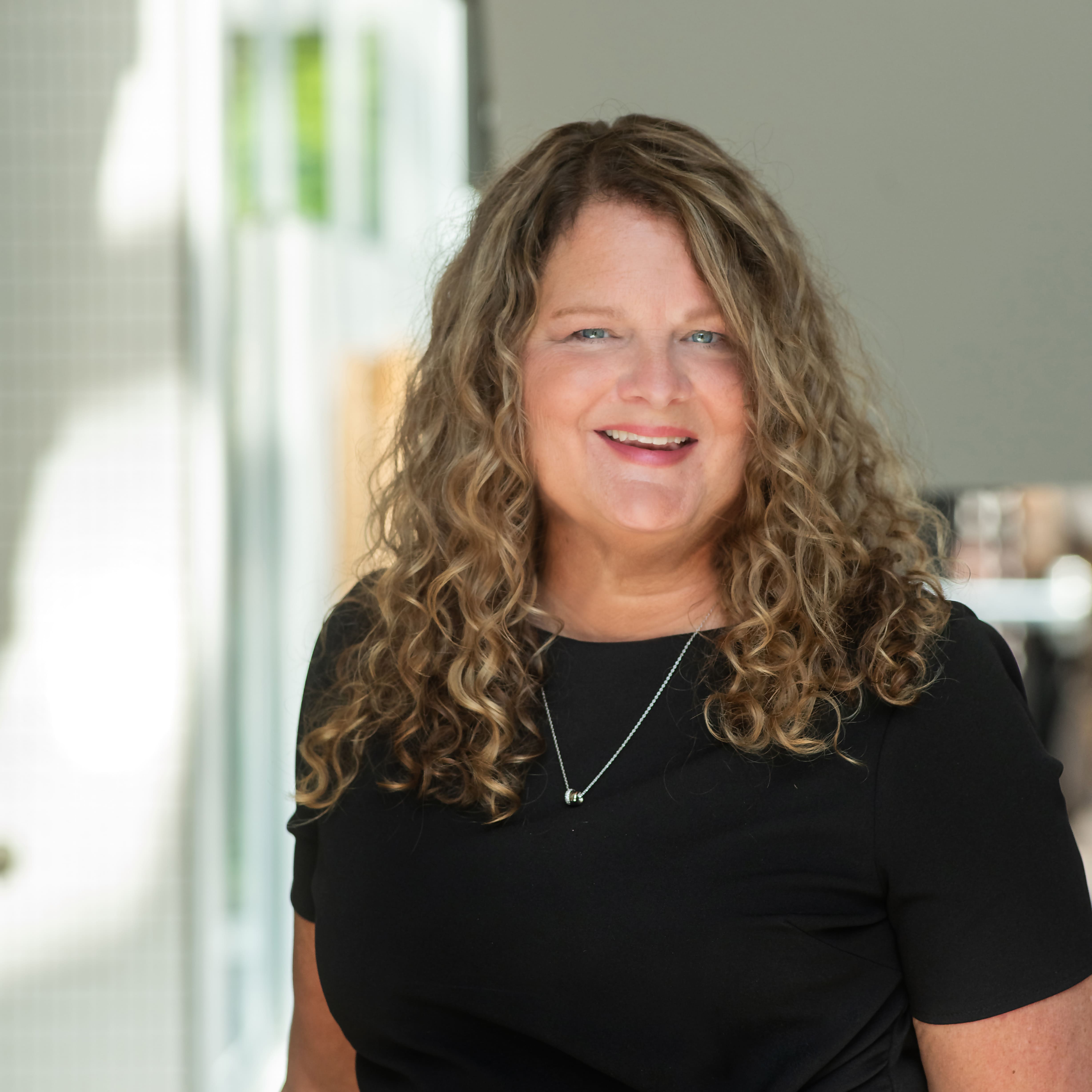 A professional headshot of Denise Thomas. She has long, curly blonde hair, is wearing a black shirt and silver necklace, and is smiling warmly.
