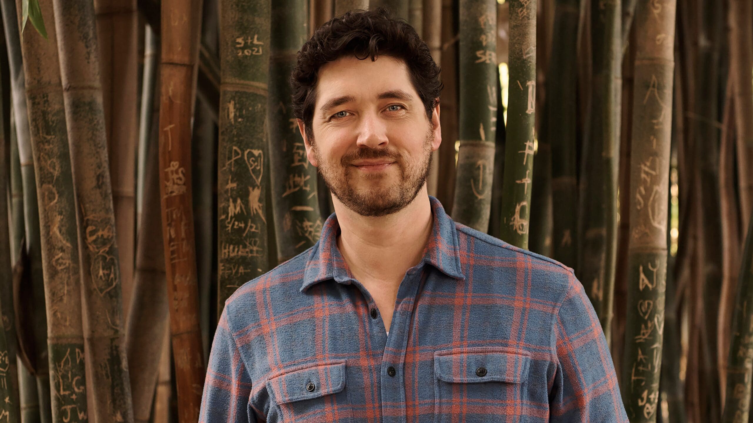 Michael Maher Jr. smiling in a blue plaid shirt, standing in a dense bamboo grove with carved stalks.