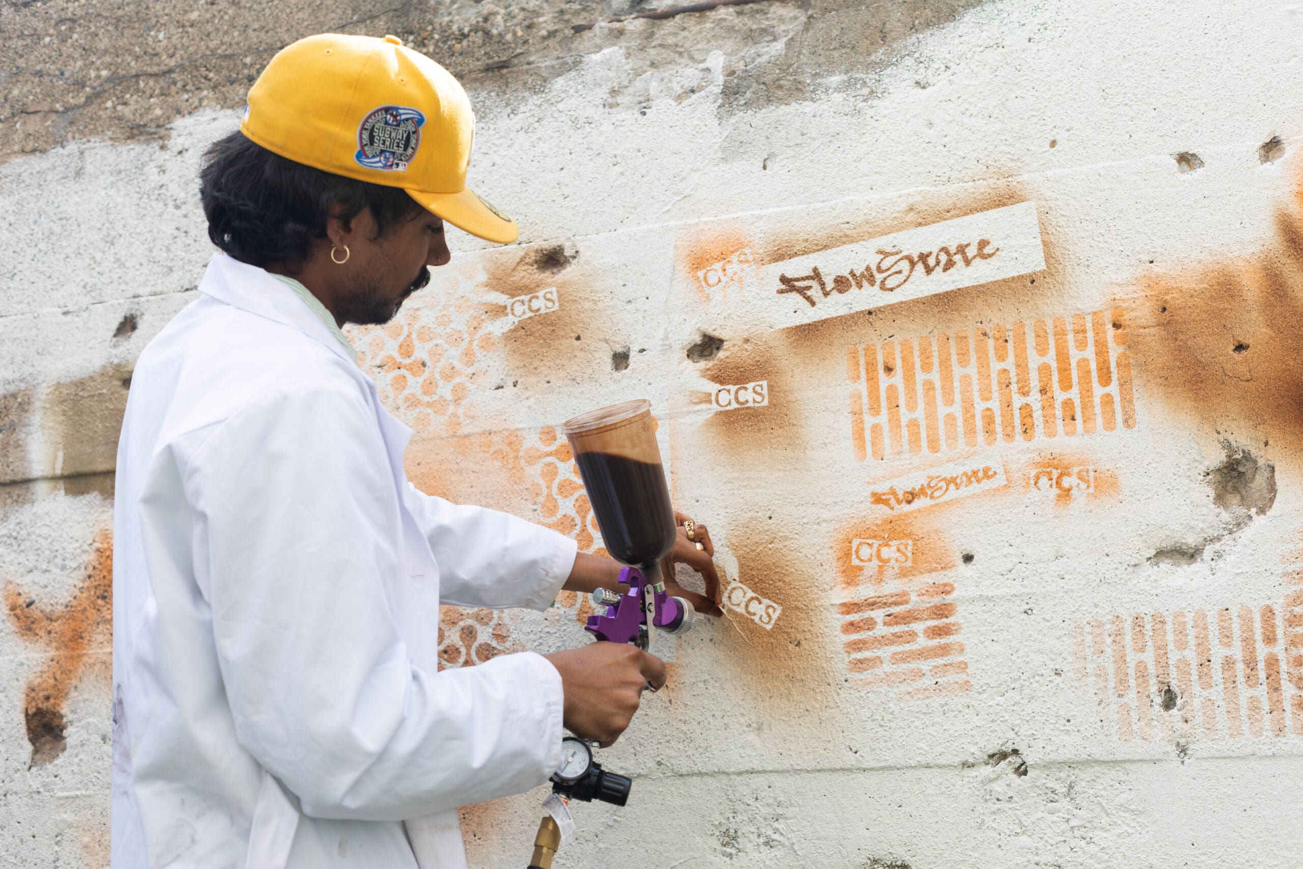 A student wearing a white lab coat, spraying bio-based sustainable spray paint on a cement wall during a project materials test at the College for Creative Studies
