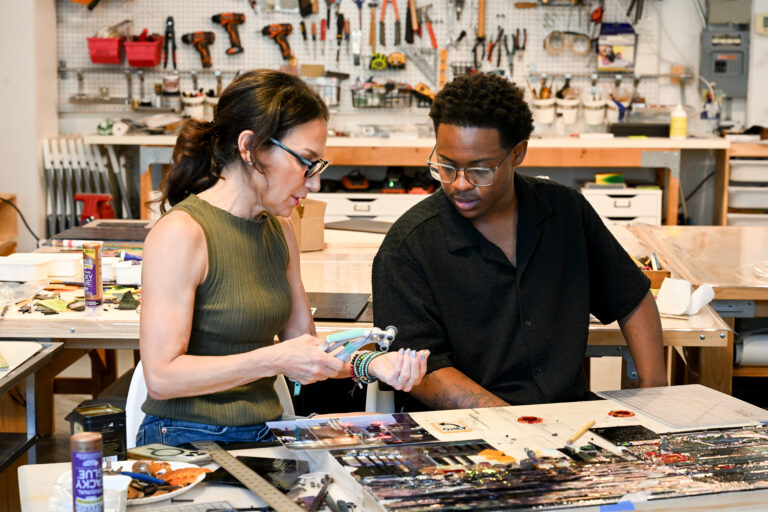 A woman and a young man wearing glasses sit together at a workbench in a craft workshop, closely examining a small hand tool and artwork.
