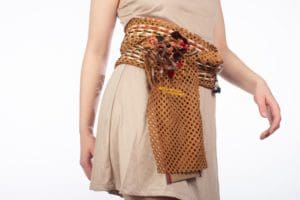 Photo of a model's torso wearing a tan tunic and a waist sash made out of dark orange perforated fabric with various types of cord and string interwoven through the holes.