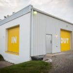 Photo of a white building with two yellow doors with white text. One door says "In" and the other says "Out"
