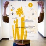 Someone holding up a white poster with black text and yellow and grey illustrations of clouds, carrots and other geometric designs