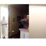 Wide shot of an elderly woman wearing a hospital gown entering from a doorway. To her left is a small tv on a table. To her right is a dark hallway and a cross hung on the wall.