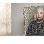 Photo of an elderly woman sitting on a ornately detailed couch. To her left is a photo of a tree in a very foggy forest.