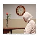 Photo of an elderly woman with her head bowed sitting at a wooden table with a single rode in a glass vase. On the white and tan wall is a large analog clock.