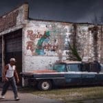 Photo of a man walking down the street in front of an old auto shop, with a faded and very chipped white mural on the side.