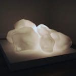 A large, bulbous cloud-like white sculpture on a display platform with lights illuminating it from underneath