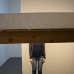 Photo of a person standing in a white room with two horizontal planks obscuring the top half of their body