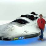 Digital rendering of a smiling man standing next to a silver futuristic concept floating vehicle with no roof and four black seat